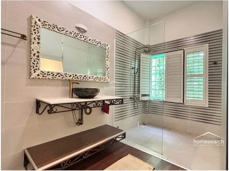 This unique bathroom showcases a blend of rustic and modern elements, highlighted by an ornate, carved white mirror frame and a vessel sink. The space includes a glass-enclosed shower area with shuttered windows for natural light and ventilation, finished with a combination of wood-look flooring and decorative wall tiles.
