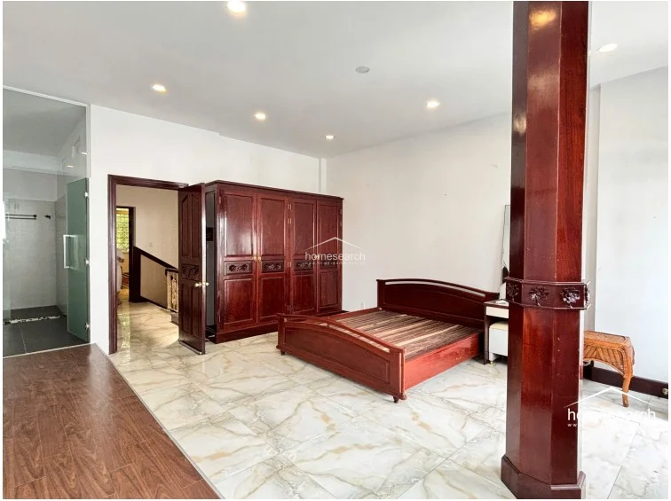 The bedroom is furnished with a substantial, traditional dark wood wardrobe that provides significant storage space, matching the wooden bed frame. The room maintains a clean, well-maintained appearance with recessed ceiling lights, marble flooring, and a clear transition into the adjacent hallway and bathroom area.
