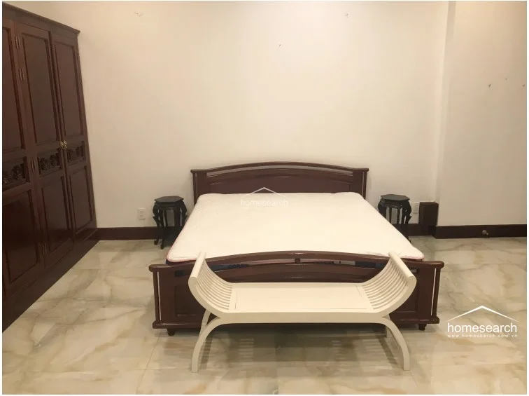This bedroom features a minimalist design with a large wooden bed frame, matching side tables, and a substantial built-in dark wood wardrobe for ample storage. The room is finished with light-colored, polished marble flooring and neutral walls, creating a bright and clean living environment.