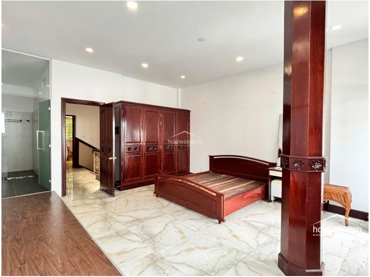 This spacious bedroom features a large, dark wood wardrobe and a matching bed frame, set against a backdrop of white walls and light-colored marble-style flooring. The room includes a private en-suite bathroom with a glass door and a large wooden support pillar that adds a traditional architectural element to the modern layout.