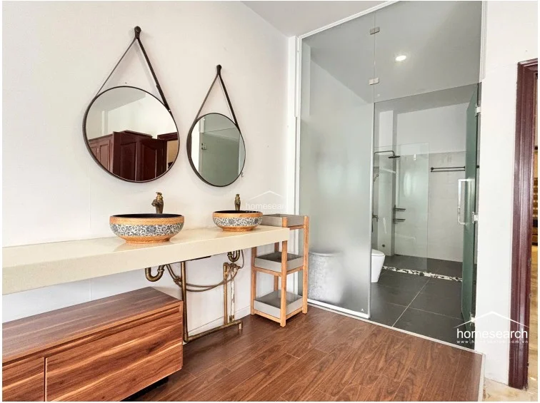 This bathroom vanity area features a unique dual-sink setup with decorative ceramic bowls mounted on a sleek stone countertop, accented by two round mirrors suspended by leather straps. The space transitions from wood-look flooring to dark tiles in the shower area, which is enclosed by a frosted glass partition for privacy.