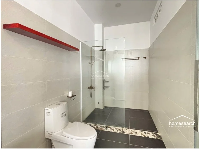 This modern bathroom is designed with a sleek, minimalist aesthetic, featuring large-format light grey wall tiles and contrasting dark floor tiles. It includes a glass-enclosed walk-in shower with a rainfall showerhead and a decorative red floating shelf, reflecting a high level of maintenance and contemporary style.
