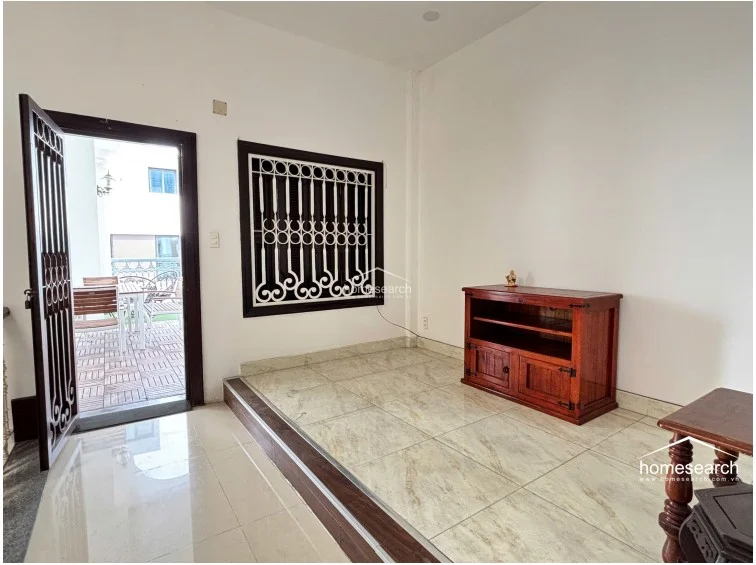 This room functions as a secondary living or lounge area, featuring a raised platform with marble-patterned tiles and a wooden media console. A door with decorative metal security bars provides direct access to an outdoor terrace, which is furnished with a table and chairs, promoting a seamless indoor-outdoor flow.