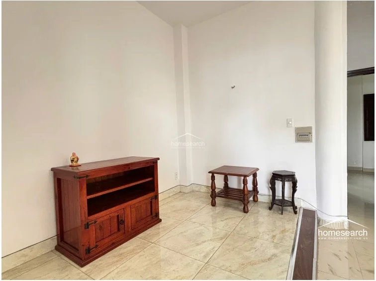 This corner of the home features a minimalist aesthetic with white walls and light-toned marble flooring, accented by a small wooden media console and traditional side tables. The area is defined by a structural column and includes a wall-mounted electrical panel, maintaining a clean and uncluttered appearance.