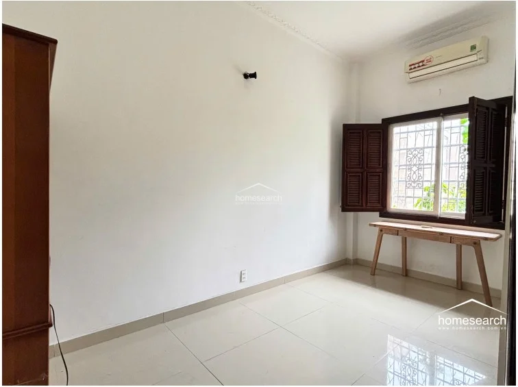 This bedroom features a minimalist layout with white walls, glossy light-colored tile flooring, and a wall-mounted air conditioning unit. A wooden desk is positioned under a window with dark wood shutters and security bars, providing functional natural light and a view of greenery.