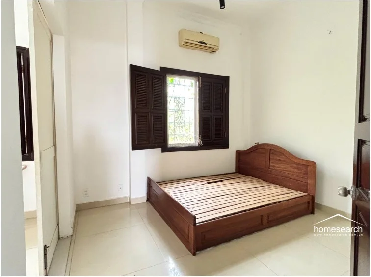 This bedroom is furnished with a solid wood bed frame and features a window with dark wooden shutters that allow for ventilation and natural light. The room is finished with white walls, light tile flooring, and a wall-mounted AC unit, maintaining a simple, well-maintained aesthetic.