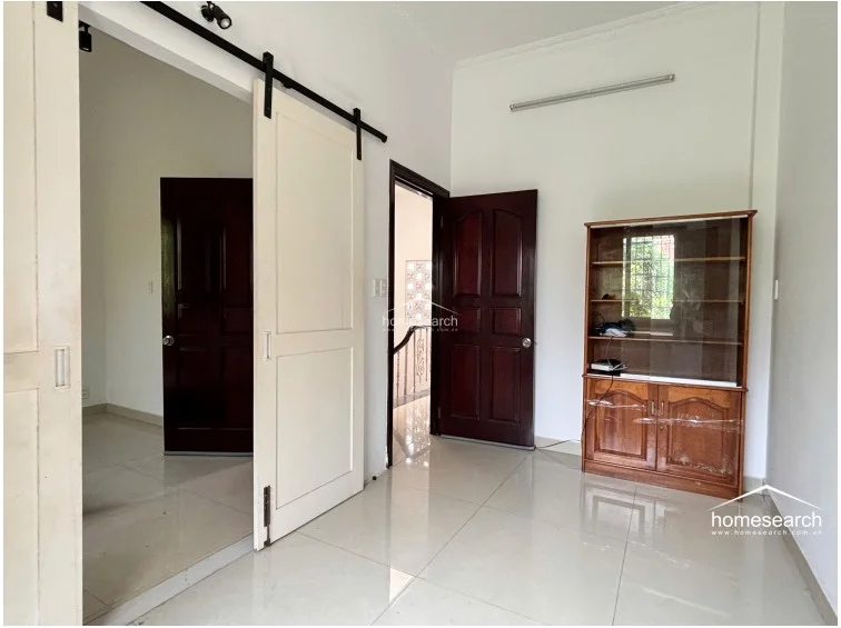 This room serves as a flexible space, featuring a sliding white barn-style door and a tall wooden display cabinet for storage. The area connects to the hallway and another bedroom, with white walls and light-colored tile flooring creating a bright, open feel.