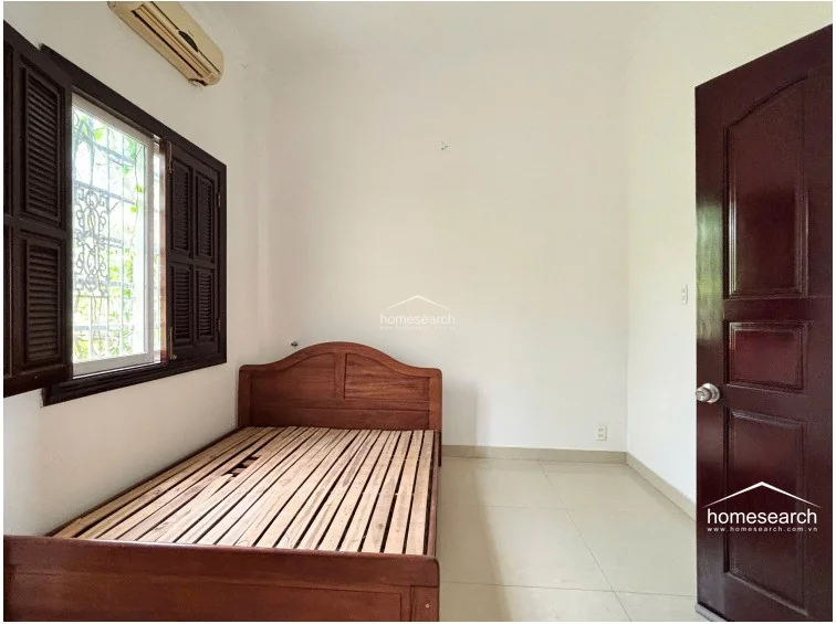 This bedroom provides a simple, functional layout with a wooden bed frame and a window equipped with dark wood shutters and security grilles. The space is finished with neutral white walls and light tile flooring, complemented by a wall-mounted AC unit for climate control.