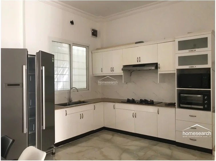 This kitchen features an L-shaped layout with white cabinetry, a dark granite countertop, and a double-basin stainless steel sink. It is well-equipped with a built-in range hood, a gas cooktop, and a large grey double-door refrigerator, all set against a clean white wall finish and light-colored tile flooring. A window above the sink provides natural light, and the space is maintained in good, functional condition.