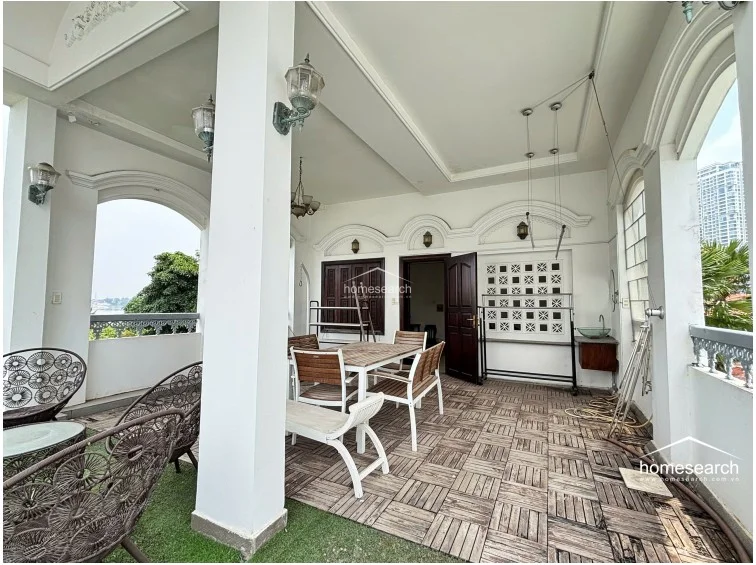 This alternate view of the terrace highlights the seamless flow between the dining area and the additional lounge space furnished with wicker chairs. The area is well-equipped with wall-mounted lighting, a small outdoor sink, and a mix of wood-tiled and artificial turf flooring, maintaining a well-kept, airy atmosphere.