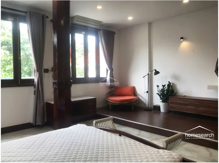 This bedroom area utilizes a raised wooden platform design, creating a distinct sleeping or lounge zone separate from the main floor. The room is brightened by two windows with grey curtains and includes a dark wood structural column, a wall-mounted AC unit, and a minimalist wooden media console. The overall aesthetic is clean and modern, with white walls and warm wood tones providing a comfortable, well-kept atmosphere.