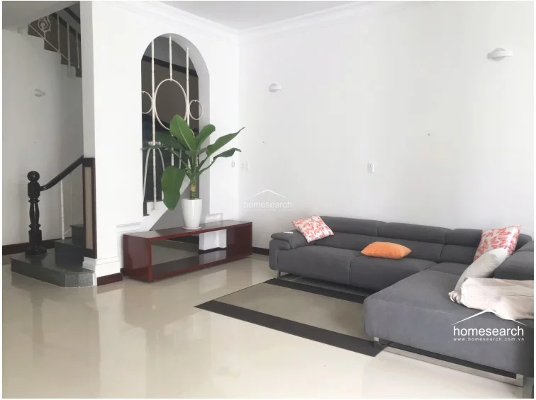 This living room showcases a spacious, open-concept layout that flows directly into the staircase area, which is highlighted by an elegant architectural archway. The room is furnished with a large grey sectional sofa and a dark wood console table, all set upon light-colored polished tile flooring. The space is bright and airy with white walls, maintaining a neutral color scheme and a high standard of cleanliness.