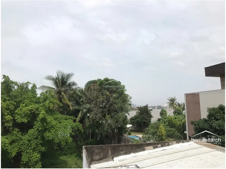 This image captures a scenic, elevated view overlooking a lush green canopy of trees and a distant body of water. The perspective suggests a peaceful, semi-rural or riverside setting with mature vegetation and a glimpse of a nearby pool. The view provides a sense of privacy and connection to nature, highlighting the property's desirable location.