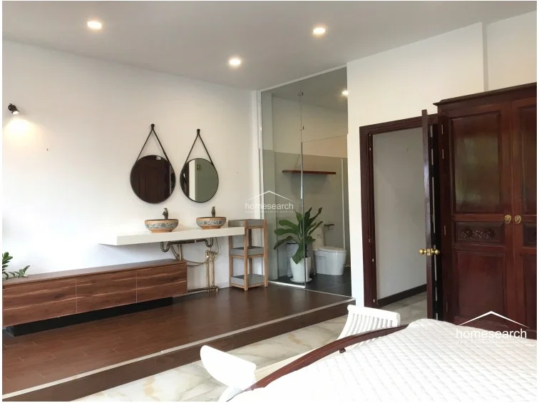 This bedroom features a unique open-concept layout with a raised wooden platform housing a dual-sink vanity area, which flows seamlessly into a glass-enclosed ensuite bathroom. The room is finished with a mix of light marble flooring in the sleeping area and rich, dark wood flooring on the platform, complemented by crisp white walls and recessed ceiling lighting. A large, ornate dark wood wardrobe provides substantial storage, while the overall aesthetic balances modern minimalist fixtures with traditional wooden elements.