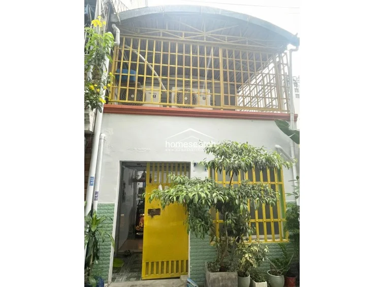 This exterior view shows a two-story residential structure featuring a yellow metal security gate and a matching protective cage over the upper-level balcony. The facade includes light-colored walls with green tiled wainscoting and a small garden area with potted plants, indicating a modest, functional maintenance level.