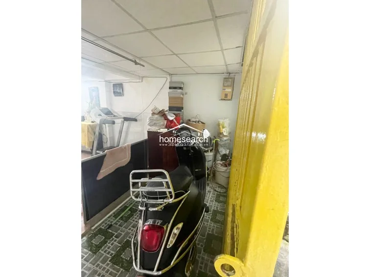 This close-up of the ground floor entryway highlights the tiled flooring and the yellow metal security gate that provides access to the interior. The space is currently utilized for indoor motorcycle parking and general storage, reflecting a practical, high-density urban layout.
