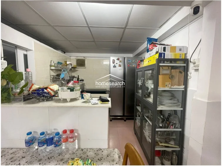 The compact kitchen area features a functional layout with a white countertop, tiled walls, and a tall stainless steel refrigerator. A dedicated glass-fronted cabinet provides organized storage for dishware, while the space connects seamlessly to the dining area, maintaining a clean and utilitarian aesthetic.