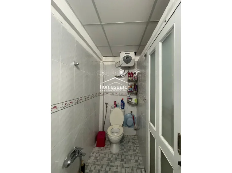 This compact bathroom utilizes a clean, white-tiled aesthetic with a decorative border and a drop-ceiling design. It is efficiently equipped with a wall-mounted water heater, a standard toilet, and a handheld shower fixture, maintaining a functional and utilitarian condition.