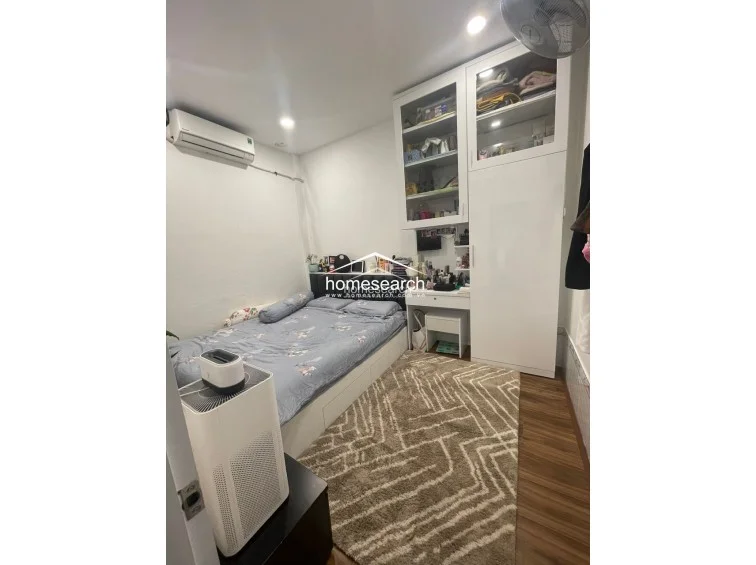 This bedroom features warm wood-look flooring, a white color scheme, and a built-in wardrobe unit that includes integrated shelving and a vanity desk. The room is equipped with a wall-mounted AC unit and a ceiling fan, creating a comfortable, well-utilized space with efficient storage solutions.