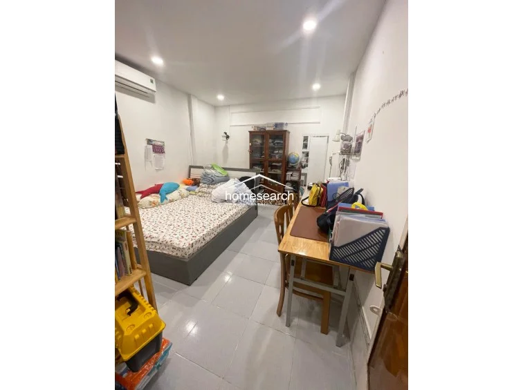 This bedroom features a functional layout with a bed positioned against the wall and a dedicated desk area for study or work. The room is finished with light-colored walls and glossy tile flooring, complemented by a wall-mounted air conditioning unit for climate control and a tall wooden cabinet for storage.