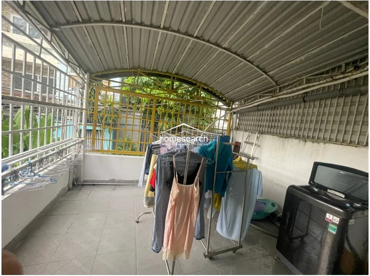 This covered outdoor terrace serves as a practical utility and laundry area, featuring a heavy-duty metal roof structure and security grilles. The space is equipped with a top-loading washing machine and ample room for drying racks, with durable tiled flooring suitable for outdoor maintenance.