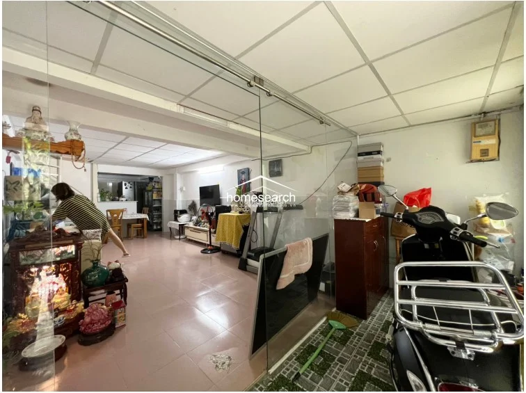 This open-plan ground floor serves as a multi-purpose living and storage area, featuring tiled flooring and a drop ceiling with recessed lighting. A large glass partition separates the front entryway from the main living space, which currently accommodates a shrine, exercise equipment, and a dining area that flows directly into the kitchen.