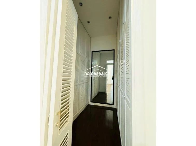 This image showcases a dedicated walk-in closet or dressing area featuring floor-to-ceiling white louvered cabinetry that provides substantial storage space. The room is finished with dark wood flooring, a full-length mirror, and recessed ceiling lighting, creating a clean, organized, and sophisticated atmosphere.