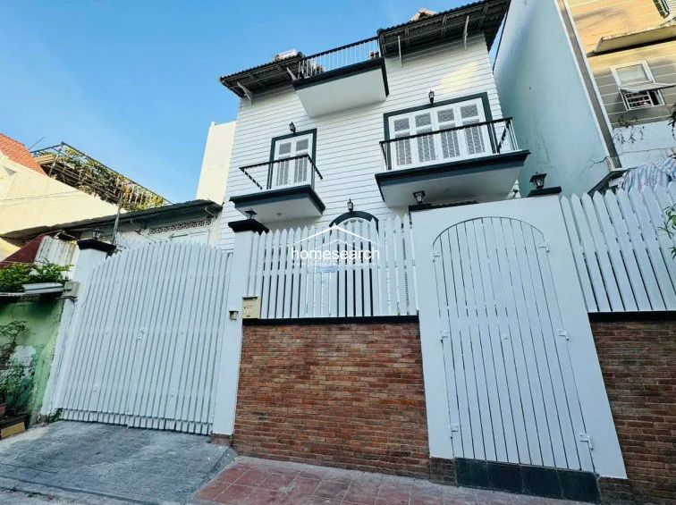 This image captures the front facade of a charming multi-level residence featuring white louvered shutters, balconies on the upper floors, and a combination of white picket fencing and brick masonry at the base. The exterior appears well-maintained with a classic aesthetic, complemented by exterior wall-mounted lighting fixtures.