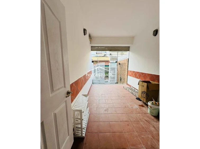This image shows a functional outdoor entryway or garage area featuring durable terracotta-style floor tiles and matching wainscoting. The space is enclosed by a white metal gate and includes wall-mounted exterior lighting, providing a secure and practical transition zone. It appears to be in good condition, offering a clean, utilitarian space for storage or vehicle parking.