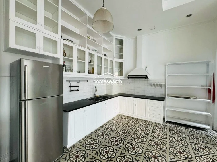 The kitchen is designed with a classic aesthetic, featuring extensive white cabinetry with glass-front doors and a contrasting black countertop. It is equipped with a stainless steel refrigerator and a built-in range hood, all set against a backdrop of decorative patterned floor tiles that add character to the workspace.