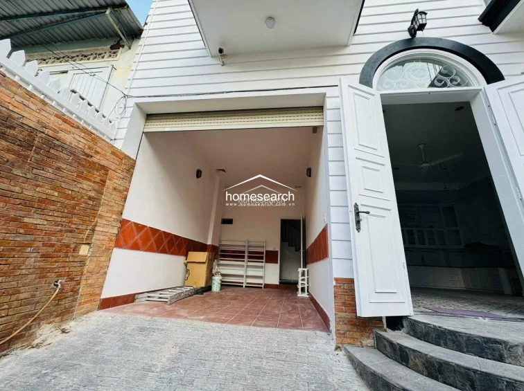 The exterior entrance features a private garage area with tiled flooring and a roll-down security door, leading directly into the home through an arched doorway. The facade combines white siding with rustic brick accents, presenting a well-maintained and secure entry point with a distinct architectural style.