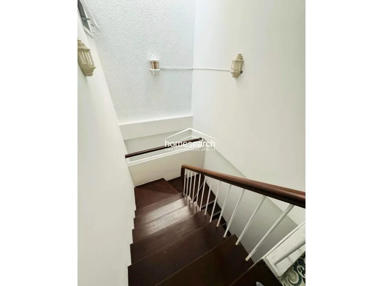 This staircase area features a classic design with dark wood treads and a polished wooden handrail supported by white balusters. The walls are finished in a clean, neutral tone, complemented by wall-mounted sconces that provide ambient lighting. The overall condition appears well-maintained, offering a bright and airy transition between levels.