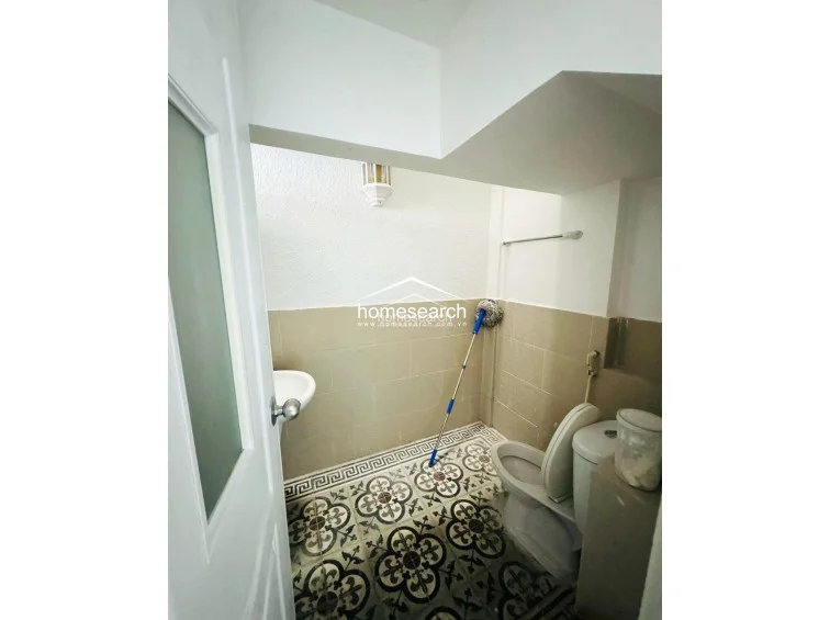 This compact bathroom features striking patterned floor tiles that add a decorative element to the functional space. It is equipped with a standard white toilet and a wall-mounted sink, with neutral beige wall tiles providing a clean backdrop. The room is well-lit and appears to be in good, hygienic condition.