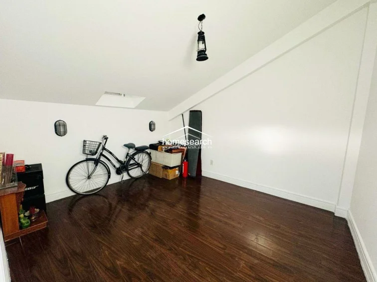 This attic-style room features sloping ceilings and dark wood flooring, providing a cozy and private atmosphere. It is illuminated by a small skylight and wall-mounted fixtures, currently serving as a storage area for a bicycle and miscellaneous items.