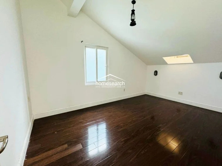 This attic-style bedroom is characterized by its dramatic sloped ceiling, which adds architectural interest to the space. The room is finished with dark wood flooring, a single window, and a skylight that allows for ample natural light, while a vintage-style pendant light serves as the primary fixture.