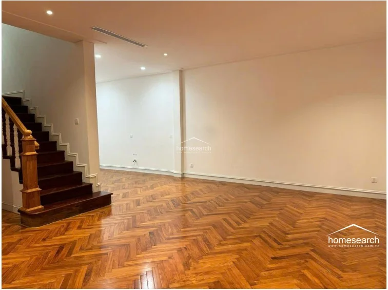 This open-plan living area features rich, herringbone-patterned wood floors and a grand wooden staircase with traditional balusters. The space is finished with clean white walls and recessed lighting, offering a versatile layout that flows naturally into the rest of the home.