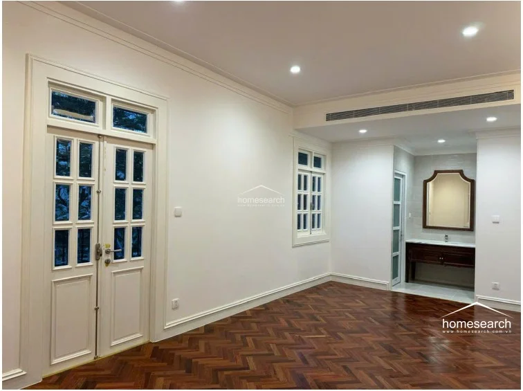 This room features elegant dark wood herringbone flooring and crisp white walls, creating a sophisticated and timeless aesthetic. The space is well-lit by recessed ceiling lights and large, multi-pane glass doors that provide a view of the exterior. A convenient vanity area with a marble countertop and dark wood cabinet is tucked into an alcove, seamlessly integrating functionality into the living space.