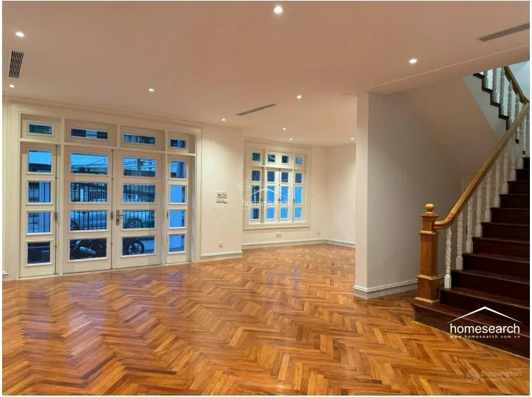 This expansive living area is characterized by its beautiful dark wood herringbone flooring and a grand staircase with a traditional wooden banister. Large white-framed glass doors and windows allow for ample natural light, while recessed ceiling lighting ensures a bright atmosphere throughout. The open layout flows seamlessly, highlighting the high ceilings and elegant architectural details of the home.
