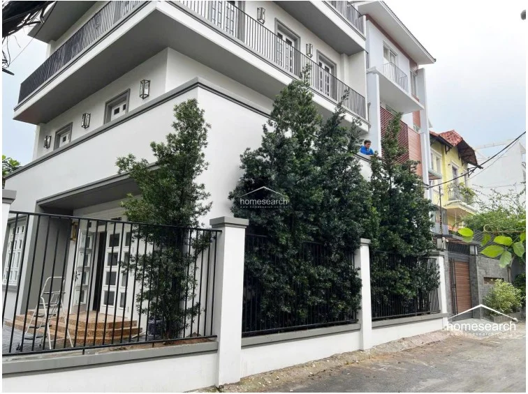 This image shows the exterior of a modern, multi-story villa featuring a clean white facade with grey trim and black metal railings. The property is well-maintained, with a gated entrance, manicured privacy hedges, and a small porch area, suggesting a high standard of external upkeep.