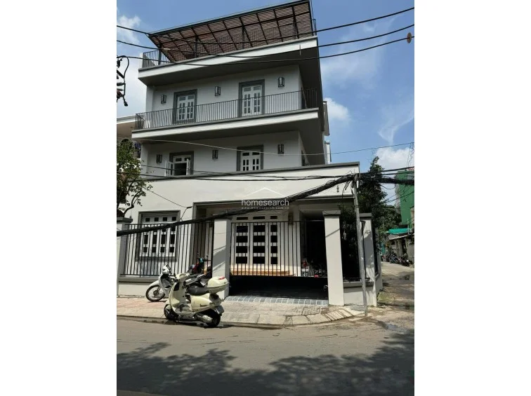 This exterior perspective highlights the side profile of the multi-story villa, emphasizing the spacious balconies and the open rooftop area with a pergola-style cover. The property is situated in an urban setting with accessible street parking and a secure gated entrance, indicating a functional and secure residential layout.