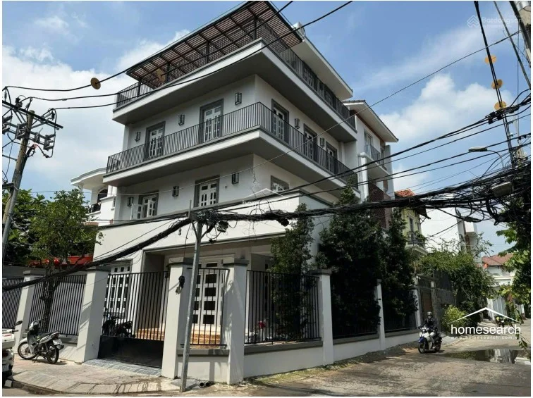 This exterior view displays a multi-story, modern-style villa with a clean white facade, multiple balconies with black metal railings, and a rooftop terrace structure. The property is secured by a perimeter wall with black metal fencing and includes a ground-level gated entrance, reflecting a well-maintained and contemporary architectural design.