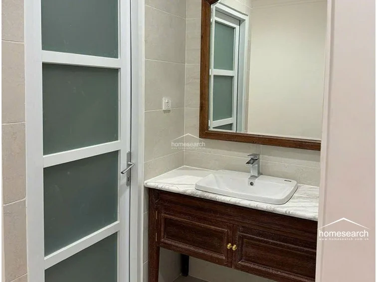 This bathroom vanity area features a dark wood cabinet with a high-quality marble countertop and a modern white basin. A large, framed mirror hangs above the vanity, reflecting the neutral-toned wall tiles and the frosted glass door leading to the rest of the bathroom. The space is clean and well-lit, reflecting a high standard of maintenance.