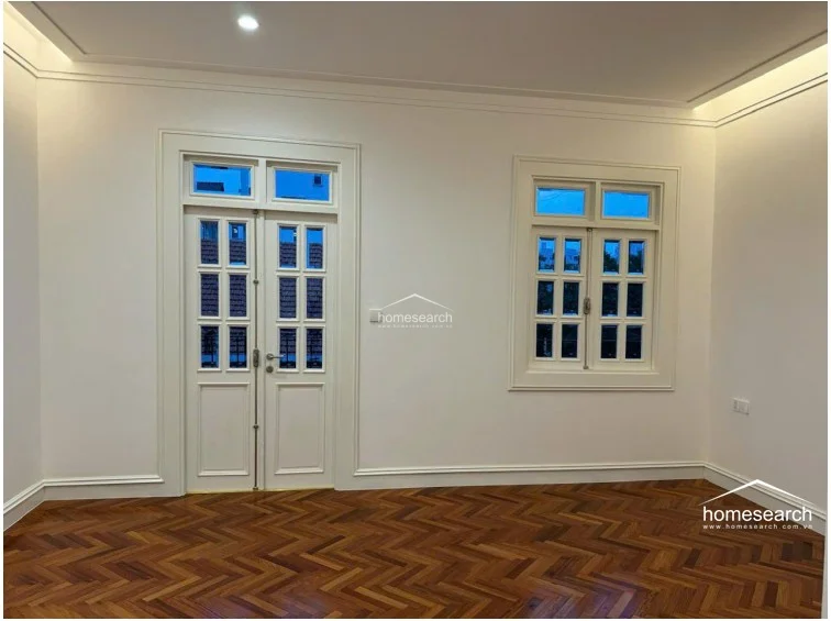 This interior space features high-quality herringbone wood flooring and crisp white walls, creating a bright and sophisticated atmosphere. The room is accented by classic white-framed French doors and matching windows that allow for natural light and a view of the neighborhood. The ceiling includes recessed lighting and subtle crown molding, indicating a well-maintained and modern finish.