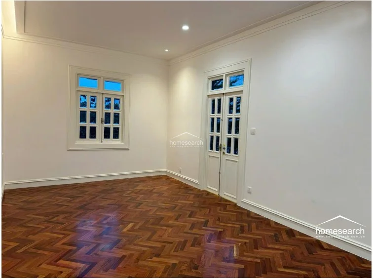 This room mirrors the design of the previous interior space, emphasizing the elegant herringbone wood flooring and bright, neutral wall palette. The symmetrical placement of the French doors and window provides a balanced architectural look, while recessed ceiling lights offer a clean, contemporary finish. The room is spacious and well-lit, presenting a versatile area suitable for a bedroom or living space.