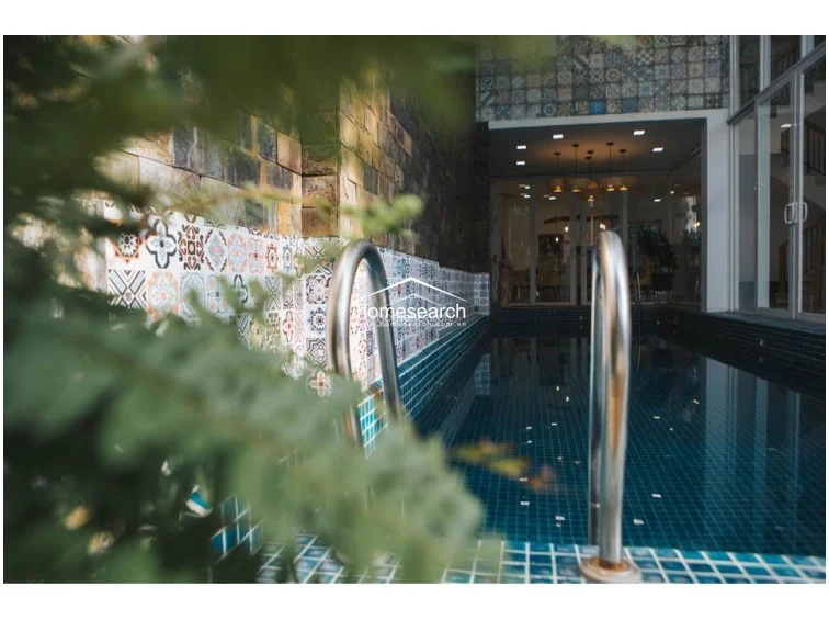 This close-up of the pool area highlights the elegant design, featuring a mosaic-tiled pool basin and a decorative accent wall adorned with intricate, patterned tiles. The space is framed by mature foliage, creating a private, serene oasis that connects seamlessly to the indoor living areas through large glass doors.