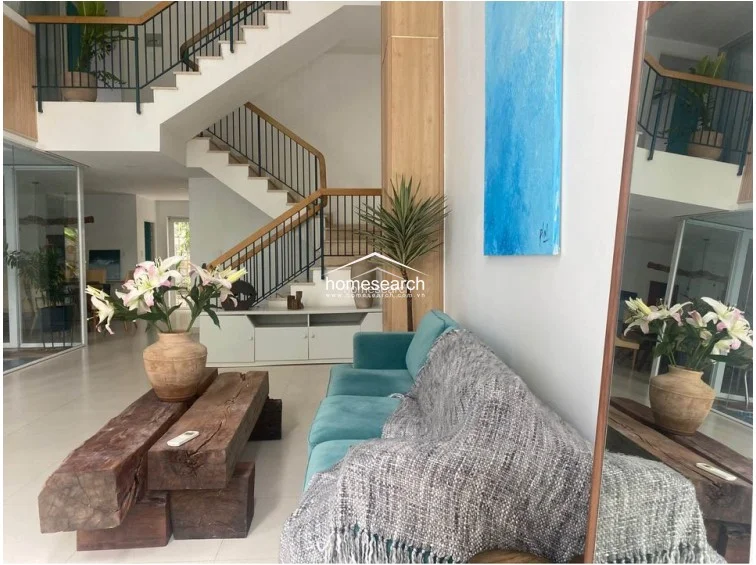 The living room showcases an open-plan layout with a double-height ceiling and a prominent staircase featuring wooden treads and a sleek black metal railing. The interior design uses a neutral color palette with light tile flooring, accented by a bold teal sofa, a rustic wooden coffee table, and large mirrors that enhance the sense of space.