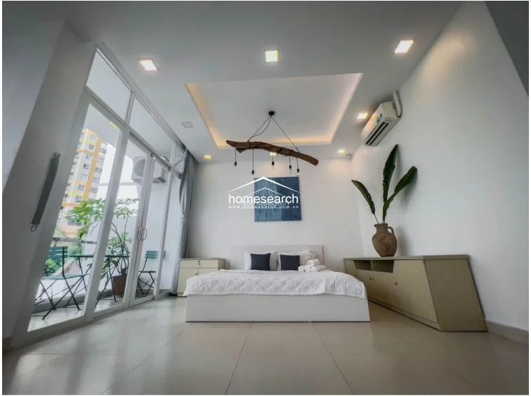 This bedroom features a minimalist design with a large glass sliding door that opens onto a private balcony, providing ample natural light and a view of the surrounding area. The room is equipped with a wall-mounted AC unit, recessed ceiling lighting, and a unique driftwood-style pendant light, all set against clean white walls and light-toned flooring.