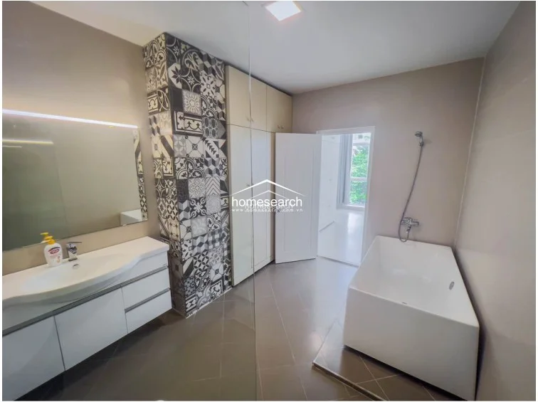The bathroom is finished with high-quality materials, including a modern white soaking tub, a sleek vanity with an integrated sink, and a striking floor-to-ceiling column decorated with monochrome patterned tiles. It includes ample storage in the form of floor-to-ceiling cabinets and maintains a bright, airy feel with neutral wall finishes and large-format floor tiles.