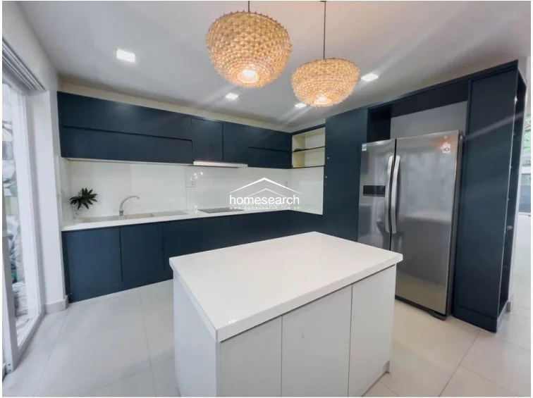 This modern kitchen features an L-shaped layout with a central island, finished in a striking matte dark blue cabinetry contrasted by crisp white countertops and backsplashes. The space is well-lit by recessed ceiling lights and two decorative woven pendant lamps, with large glass sliding doors providing natural light and direct access to an outdoor area. The flooring consists of large-format light-colored tiles, and the kitchen is equipped with a stainless steel refrigerator and integrated appliances, presenting a clean, well-maintained contemporary aesthetic.