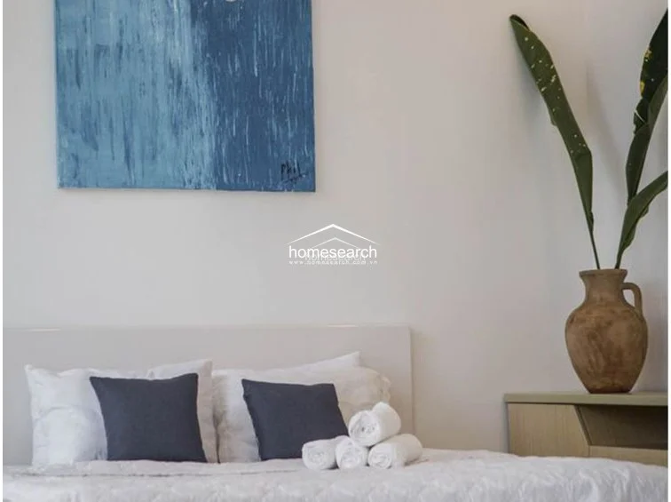 This bedroom features a minimalist, serene design with neutral white walls accented by a large, textured blue abstract painting and a decorative plant in a rustic clay vase. The bed is dressed in clean white linens with dark blue accent pillows that coordinate with the home's color scheme, and the room includes a simple light-wood bedside cabinet. The overall atmosphere is calm and well-maintained, focusing on a clutter-free, boutique-style presentation.